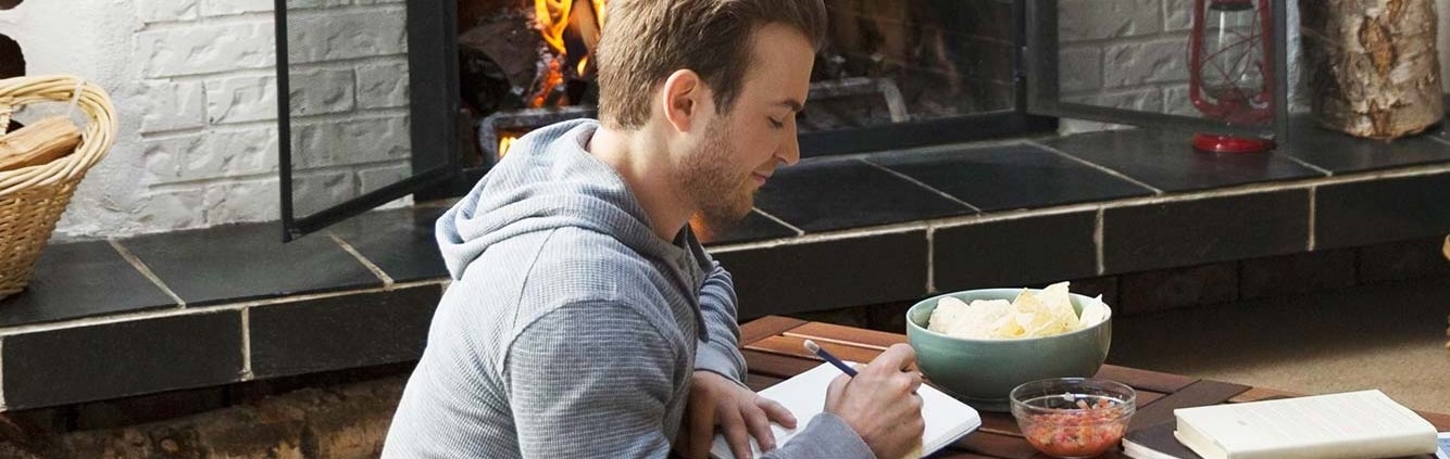 Man writes in a journal on a table with a fireplace in the background.