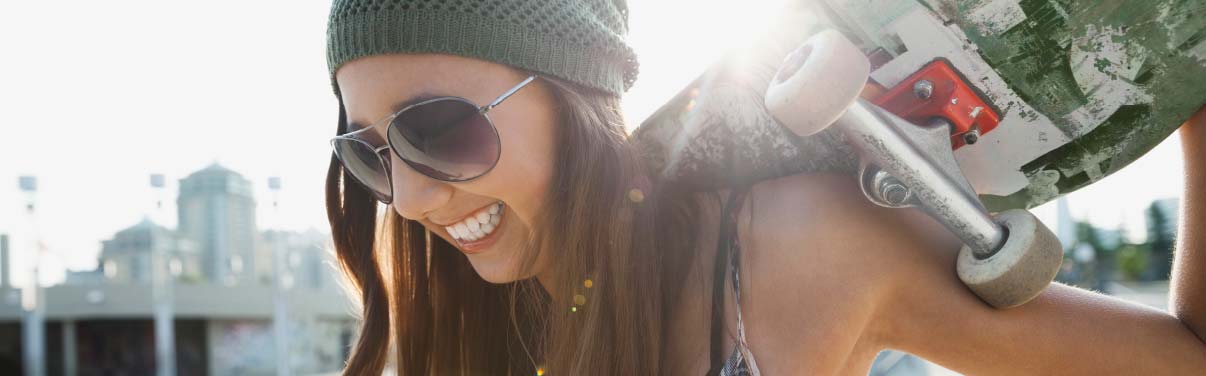 A smiling teenage girl wearing a beanie and sunglasses and holding a skateboard
